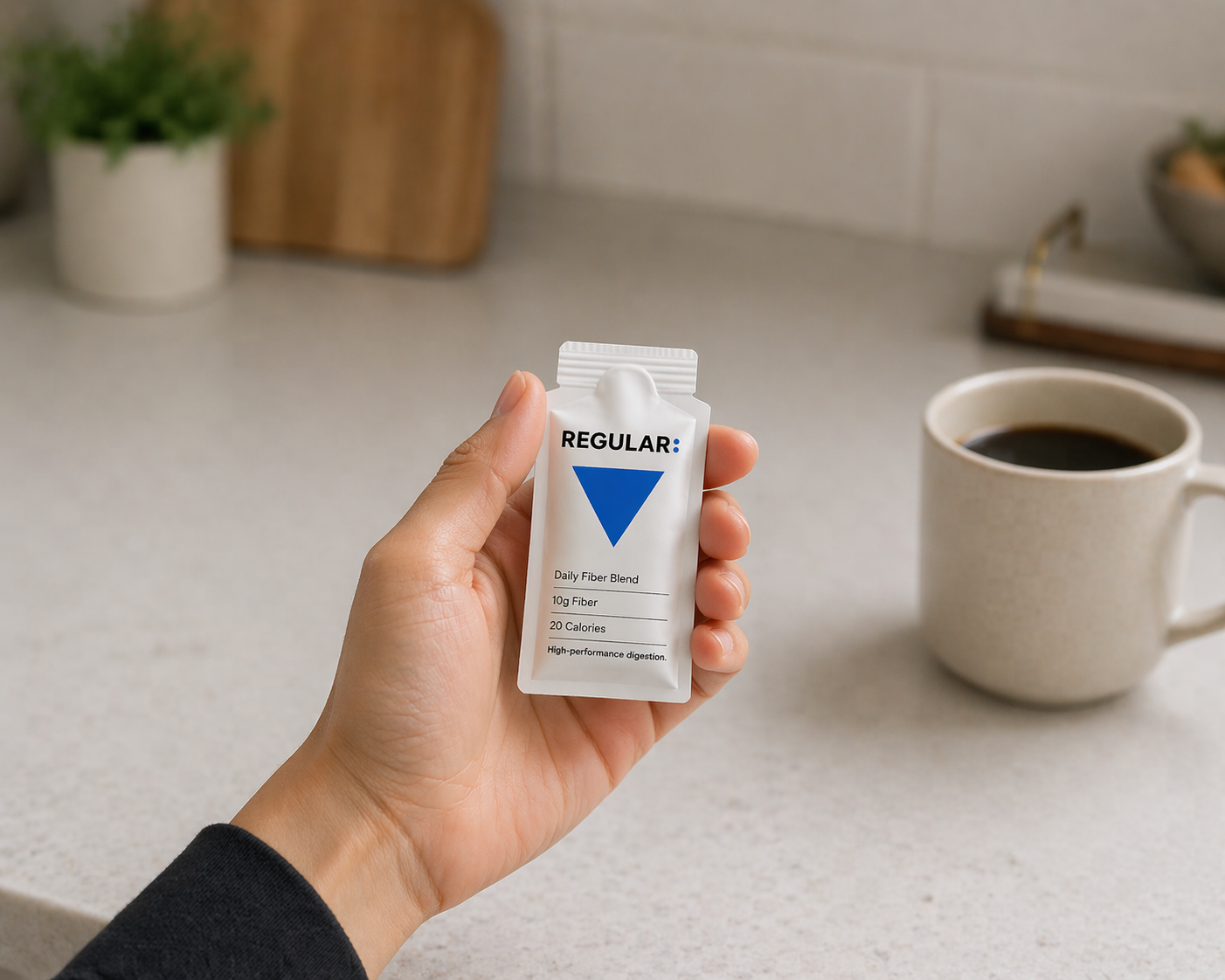 Hand holding a fiber supplement packet with a coffee cup in the background on a kitchen counter.
